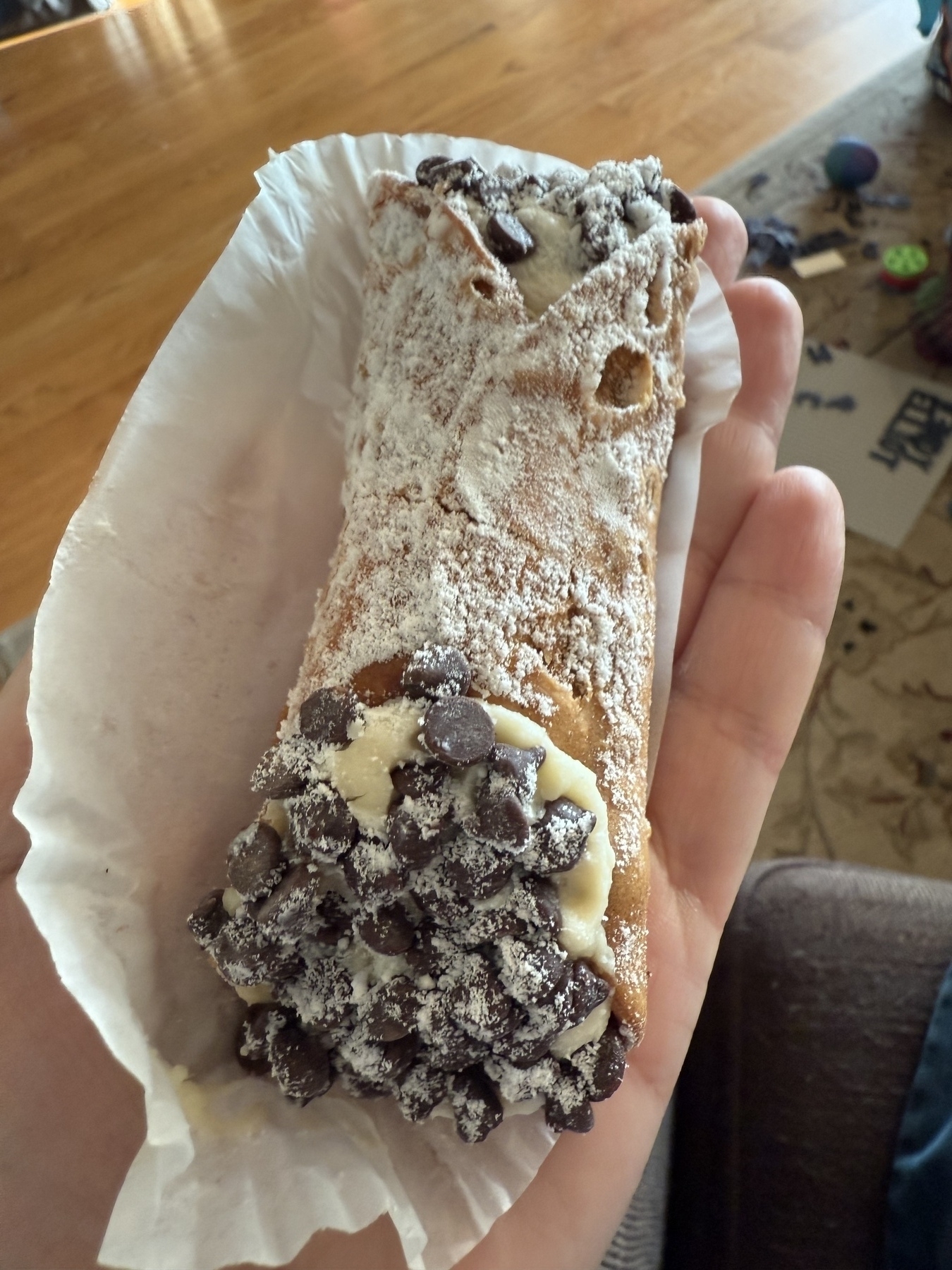 A hand is holding a cannoli filled with cream and topped with chocolate chips, dusted with powdered sugar, resting on a paper liner.