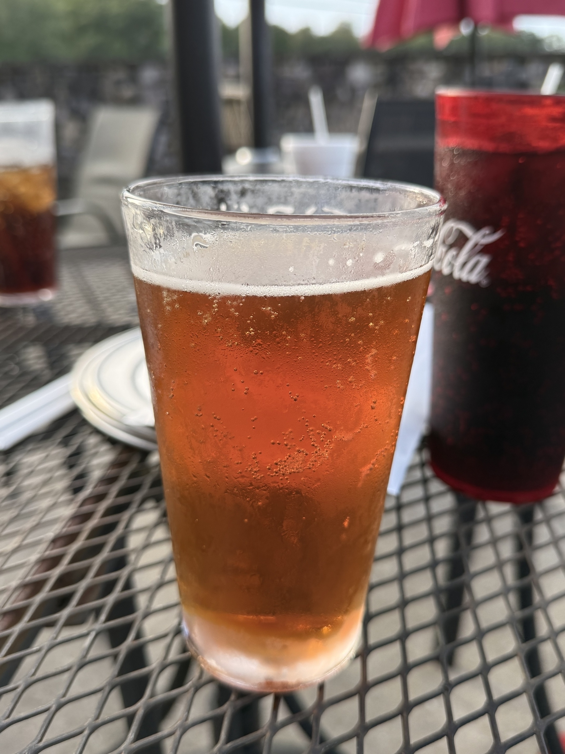 A mostly full glass of amber-colored beer is on a metal patio table alongside a red cup and other dining items.