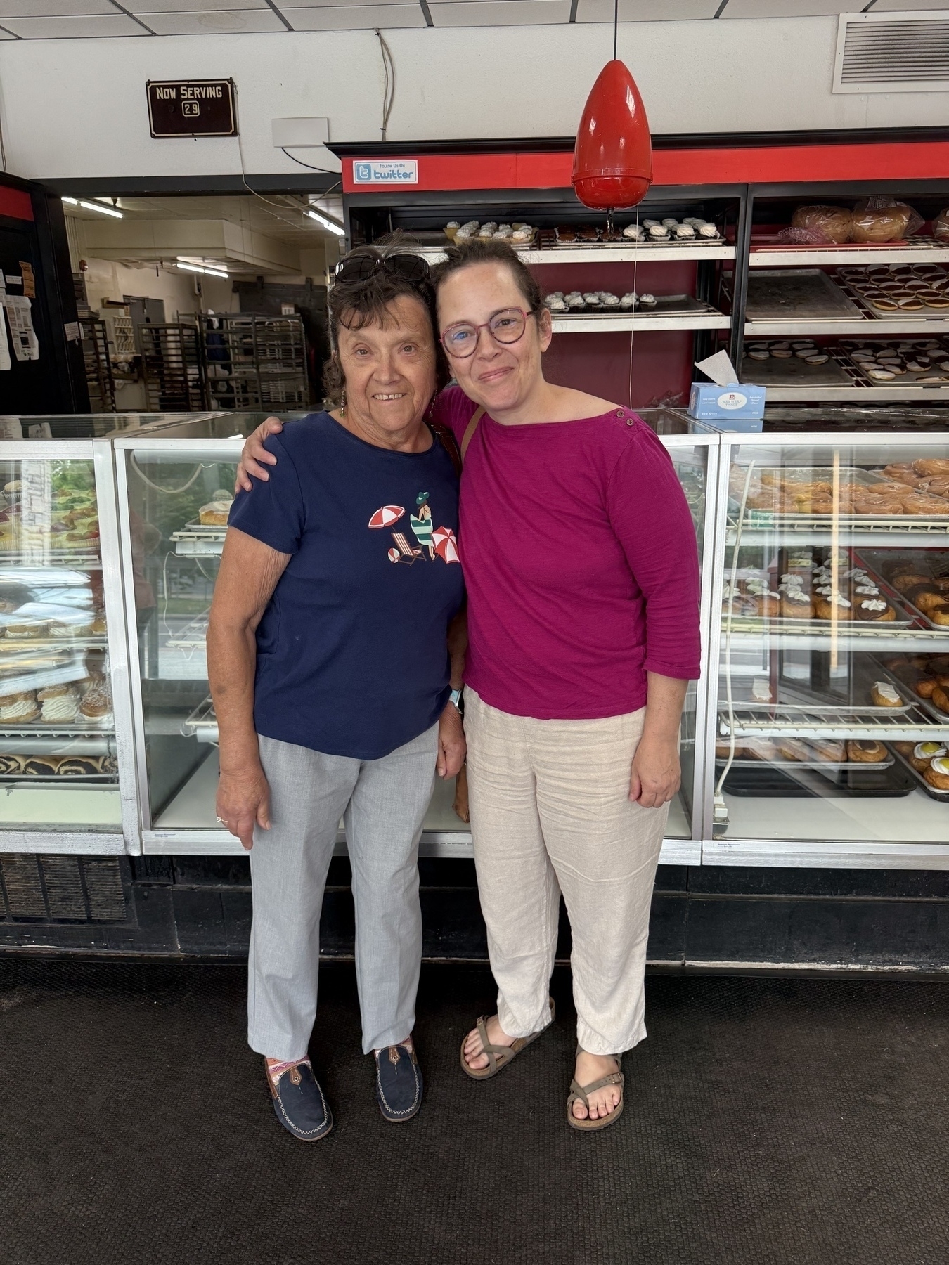 Two women are standing in front of a bakery display case filled with pastries.
