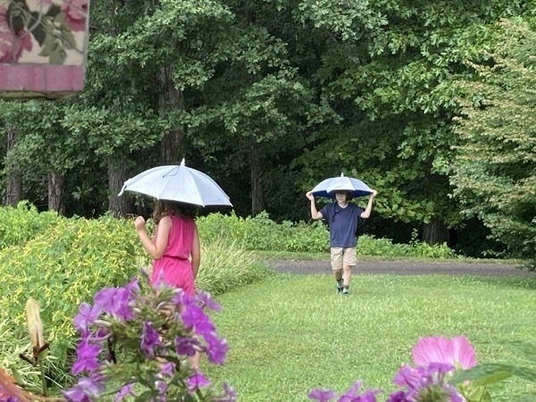 Two children playing on a green lawn surrounded by flowers and trees. It is raining, and both are wearing umbrella hats. 